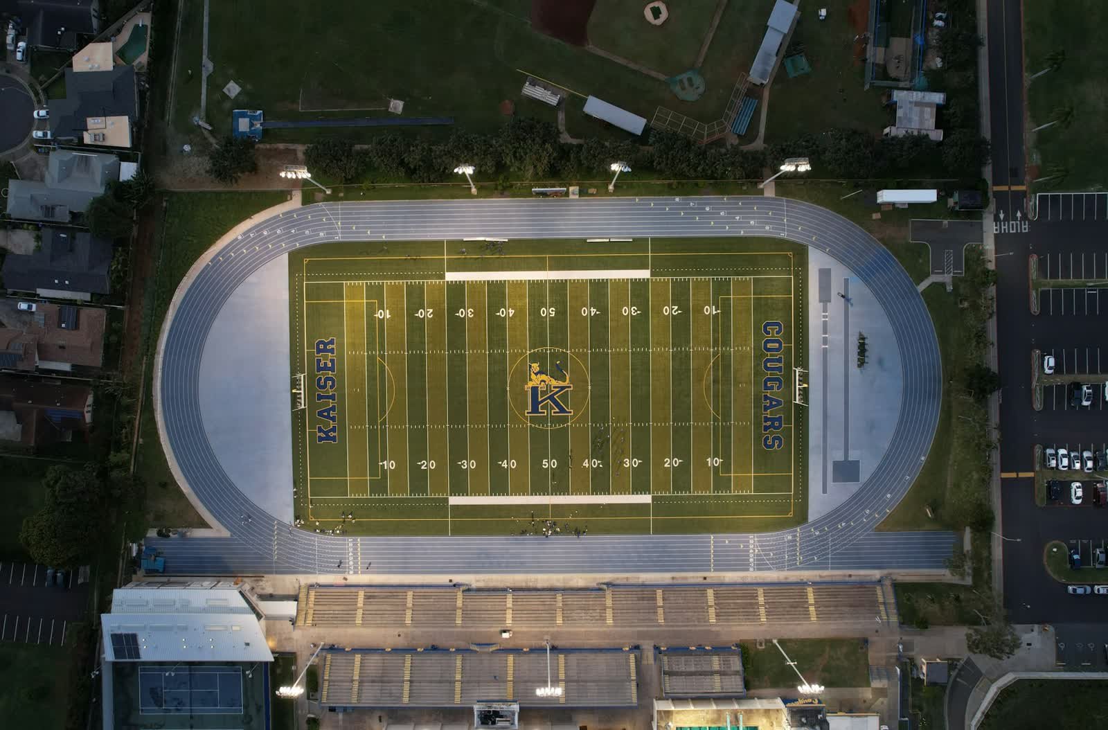 Aerial view of a lit American football field with yard lines, surrounded by a running track and bleachers