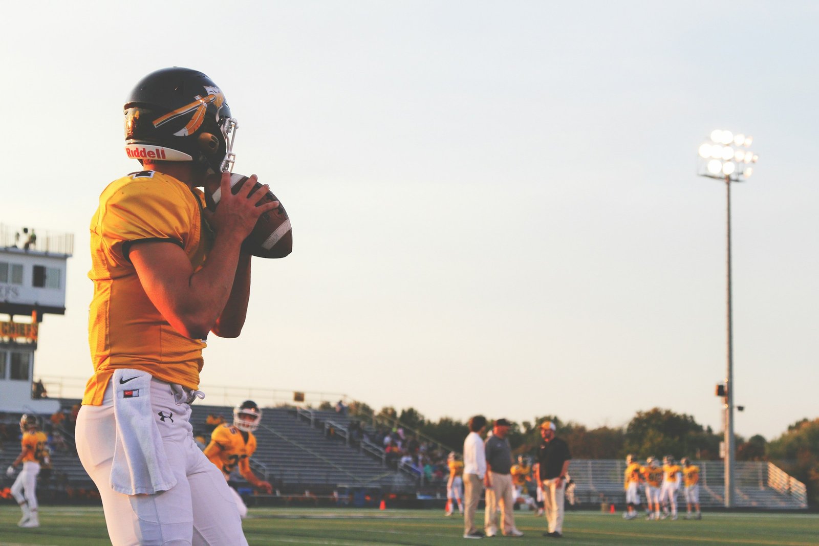 American football player holding a brown oval football under stadium lights